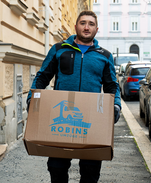 Eine Person mit einem großen Karton mit der Aufschrift „ROBINS UMZÜGE“ läuft eine schmale, von parkenden Autos gesäumte Stadtstraße entlang und unterstreicht die Effizienz des Umzugsservice.