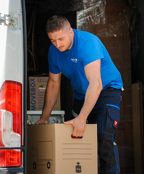 Ein Mann im blauen Hemd lädt einen braunen Karton auf die Ladefläche eines Lastwagens. Der Karton, beschriftet mit einem LKW-Symbol und „30 kg“, scheint Teil eines effizienten Lieferservices für Geschäftskunden zu sein.
