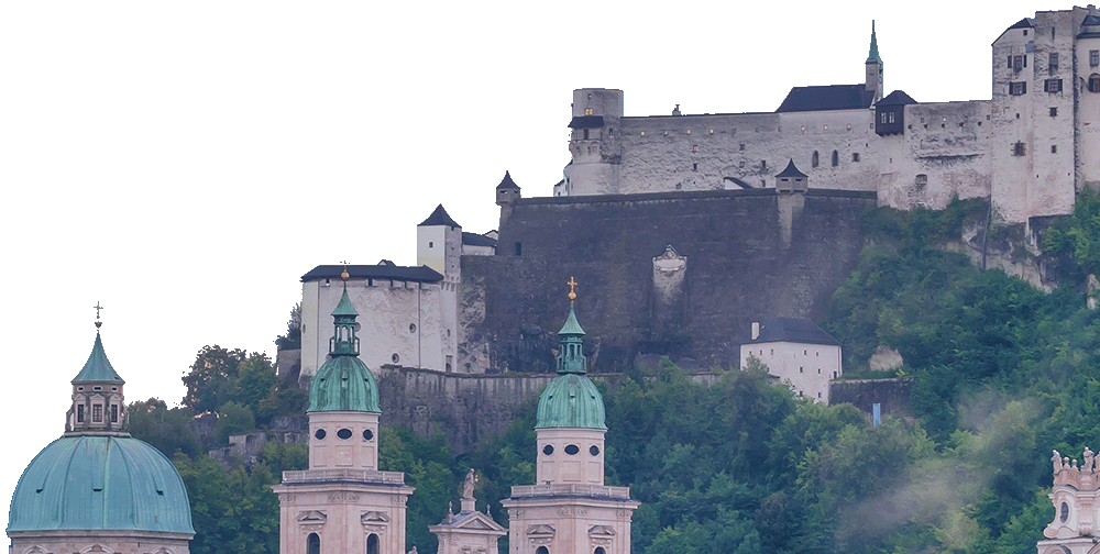 Auf einem Hügel thront eine Burg mit hoch aufragenden Steinmauern und Türmchen, die im Vordergrund eine Kirche mit grünen Kuppeln überblickt. Teile des Hügels sind von dichtem Grün bedeckt.