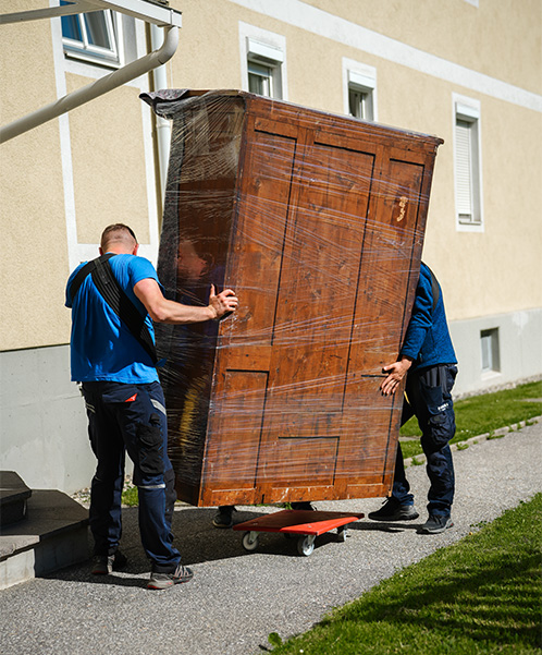 Zwei Männer bewegen einen großen, in Plastik eingewickelten Holzkleiderschrank auf einem Rollwagen vor einem Gebäude.
