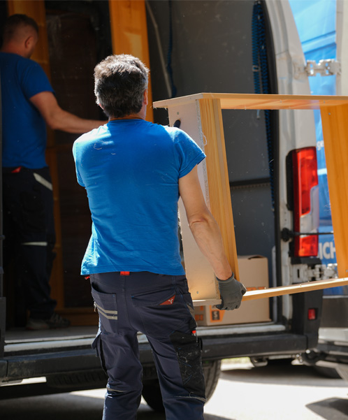 Zwei Arbeiter in blauen Hemden laden Holzmöbel in einen weißen Lieferwagen. Einer der Arbeiter ist teilweise im Lieferwagen zu sehen, der andere trägt ein Möbelstück.