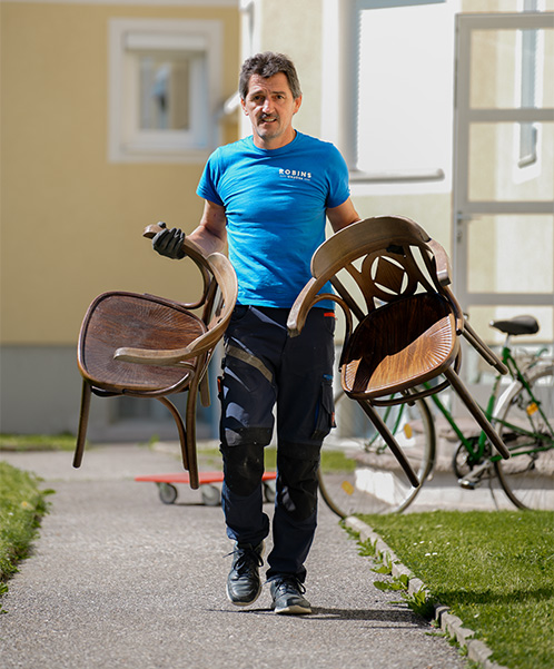 Ein Mann im blauen Hemd trägt zwei Holzstühle nach draußen. Im Hintergrund sind ein Gebäude, ein Fahrrad und eine rote Transportkarre zu sehen.