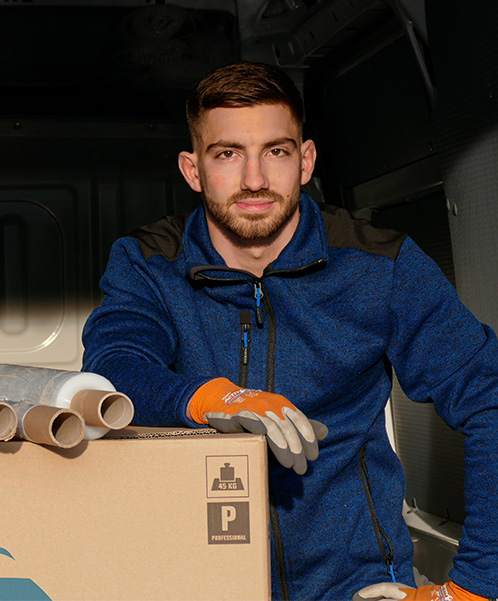 Ein Mann in blauer Jacke und orangefarbenen Handschuhen steht in einem Lieferwagen und lehnt sich an einen großen Karton. Neben ihm sind Rollen aus Papier oder Stoff zu sehen.