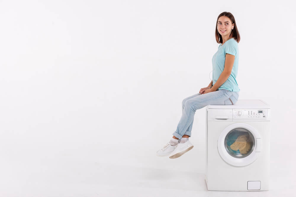 Eine Frau in blauem Hemd und Jeans sitzt vor weißem Hintergrund an einer Frontlader-Waschmaschine.