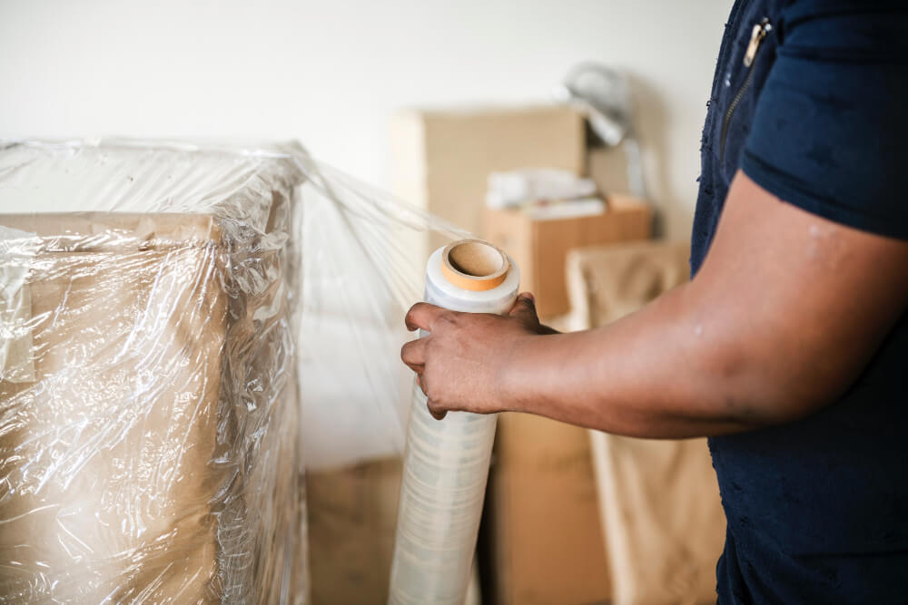 Eine Person verpackt ein großes Paket in Plastikfolie und steht neben Kartons.