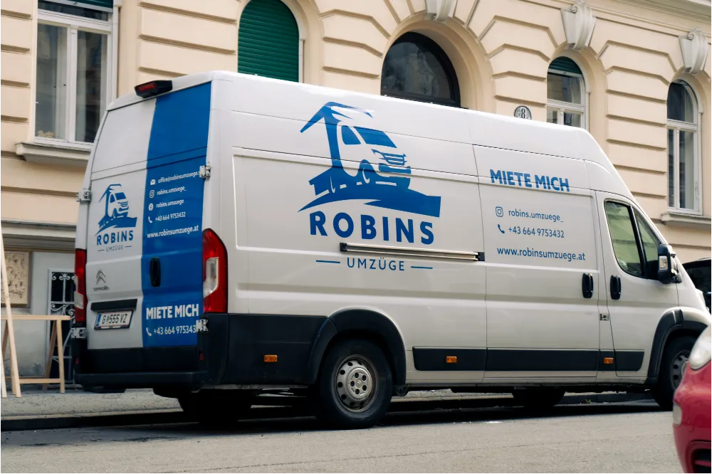 Ein weißer Umzugswagen mit blauem „Robins Umzüge“-Branding und Kontaktinformationen, der eine effiziente und kostengünstige Umzugsfirma-Kostenlösung darstellt, ist auf einer Straße vor einem beigen Gebäude geparkt.