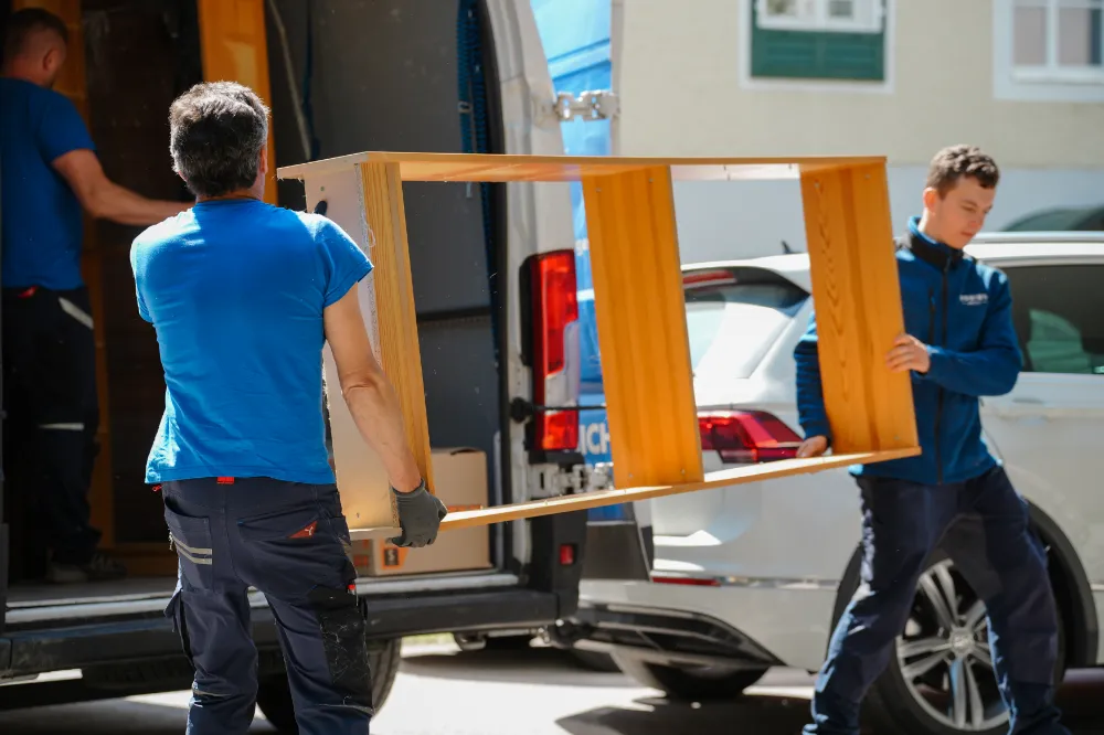 Zwei Männer in blauen Hemden sind gerade dabei, im Freien geschickt ein Holzregal aus einem Lieferwagen zu transportieren. Dabei demonstrieren sie die Effizienz und Wertschöpfung, die oft mit der Umzugsfirma „Kosten“ in Verbindung gebracht werden.