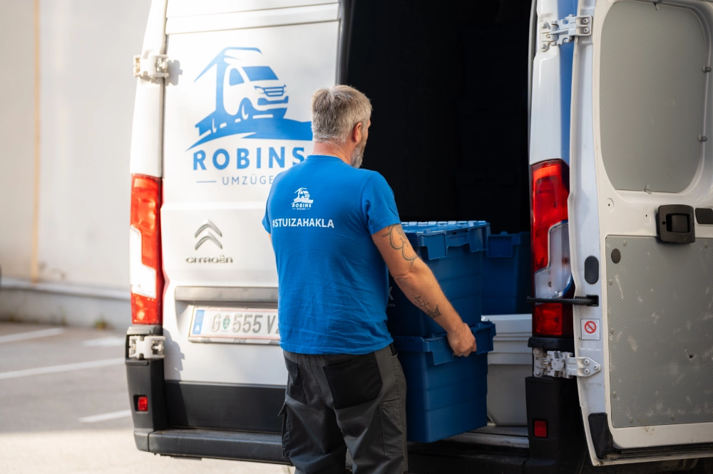 Eine Person im blauen Hemd lädt blaue Kisten in einen weißen Lieferwagen mit dem „ROBINS UMZÜGE“-Logo und bereitet sich auf einen Umzug von Österreich nach Deutschland vor.