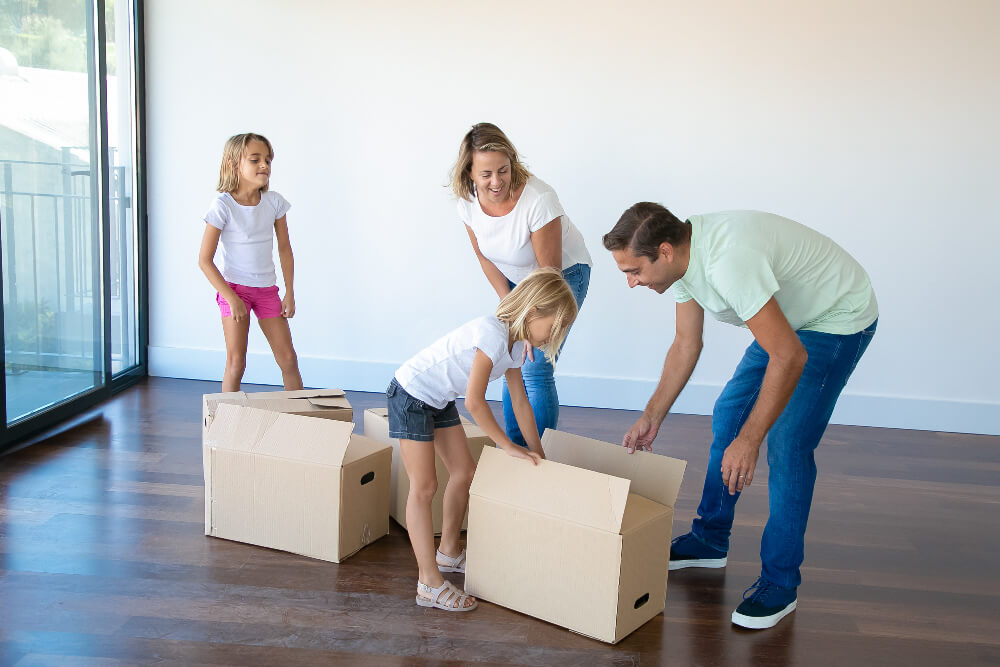 Eine Familie mit zwei Kindern öffnet Kartons in einem unmöblierten Raum mit Holzboden und großen Fenstern und markiert damit den Beginn ihres Umzugs von Österreich nach Deutschland.