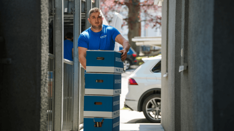 Eine Person im blauen Hemd, die wahrscheinlich darüber nachdenkt, wie sie einen effizienten Umzug planen könnte, trägt vier gestapelte Kisten nach draußen in die Nähe eines grauen Gebäudes und eines weißen Autos.