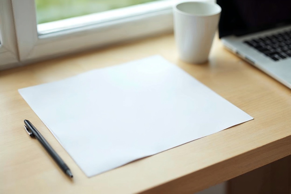 Schreibtisch mit geschlossenem Laptop, leerem Blatt Papier und Stift bei Tageslicht